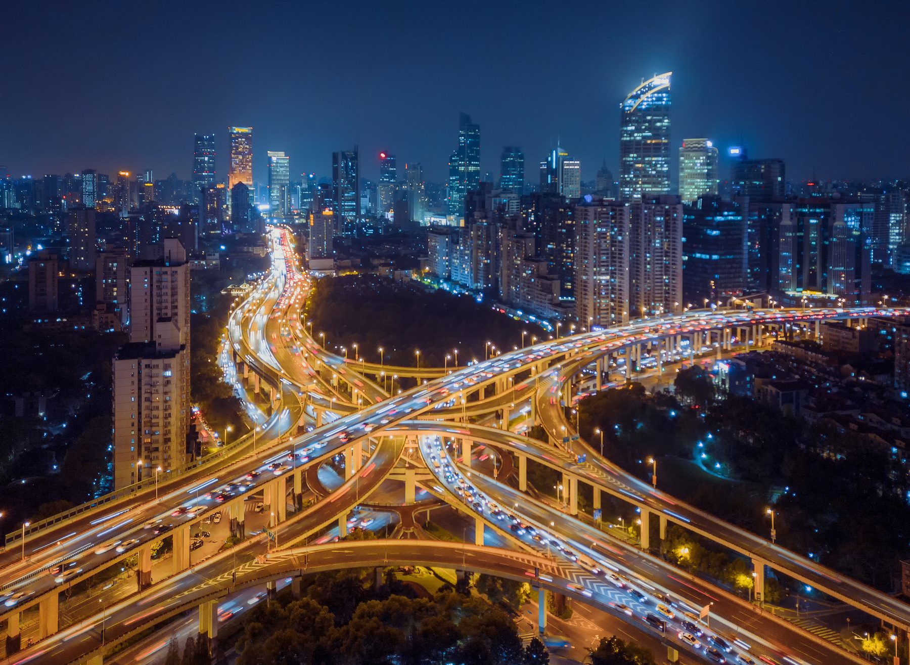 A multi-level urban interchange at night, lit along every deck, with the city skyline behind